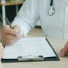 Medic in halat alb completand documente medicale la birou, alaturi de pacient