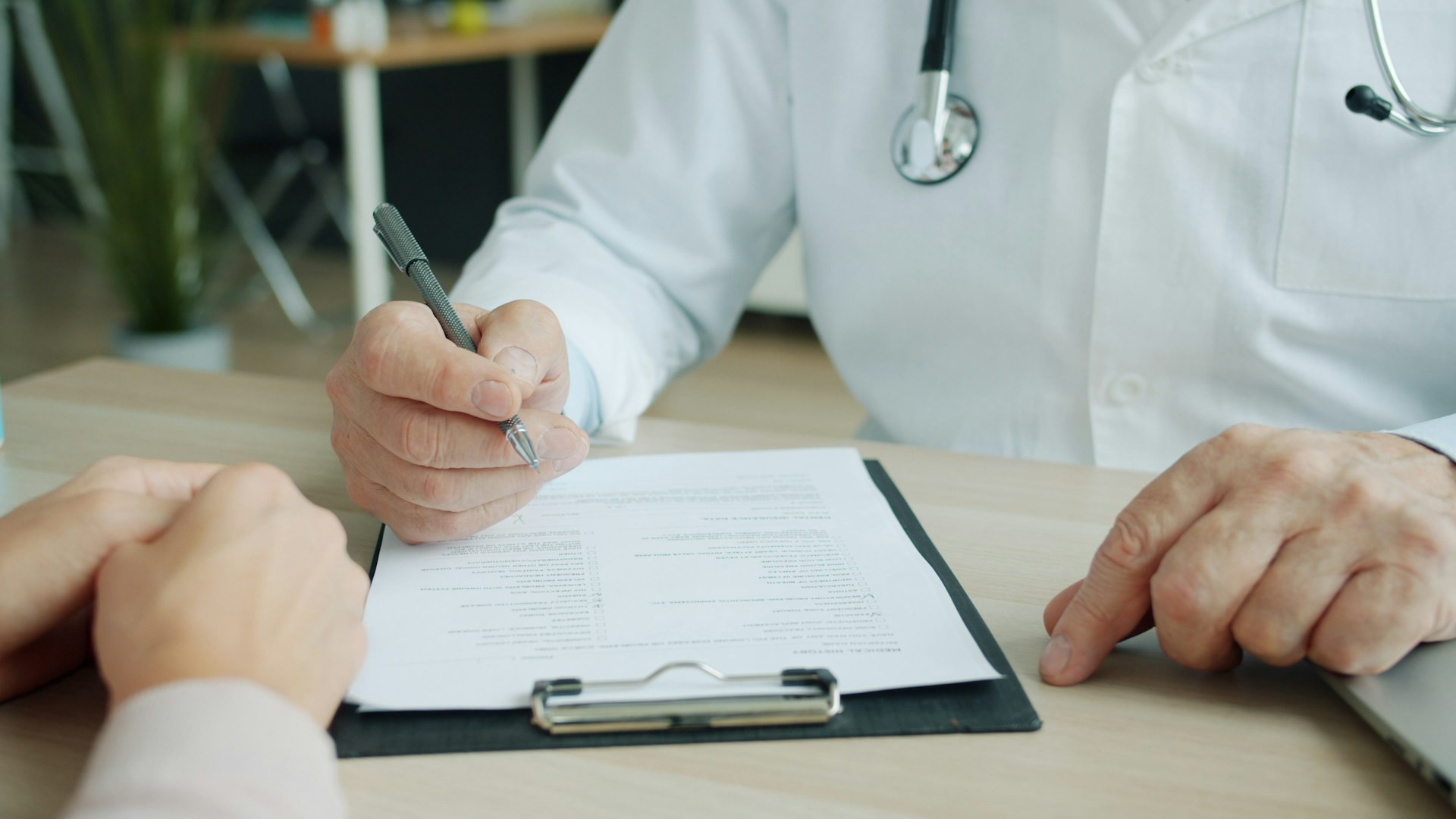 Medic in halat alb completand documente medicale la birou, alaturi de pacient
